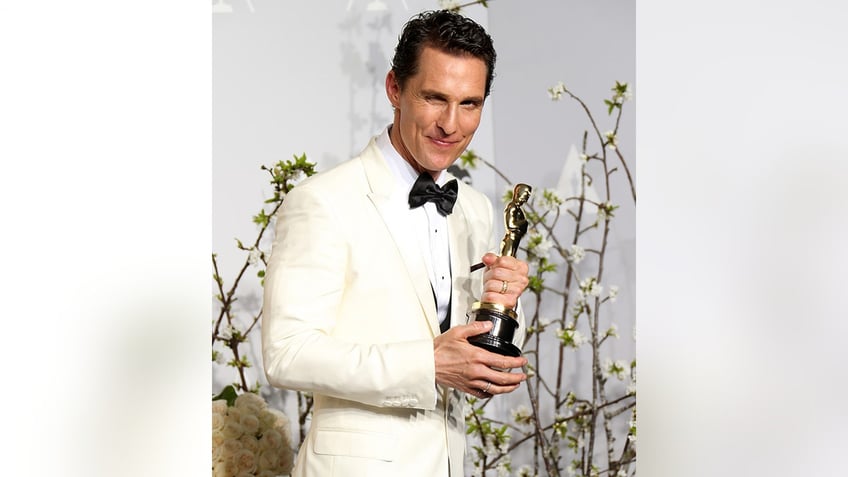 Matthew McConaughey in an off-white tuxedo winks and looks to his right holding his Oscar Award