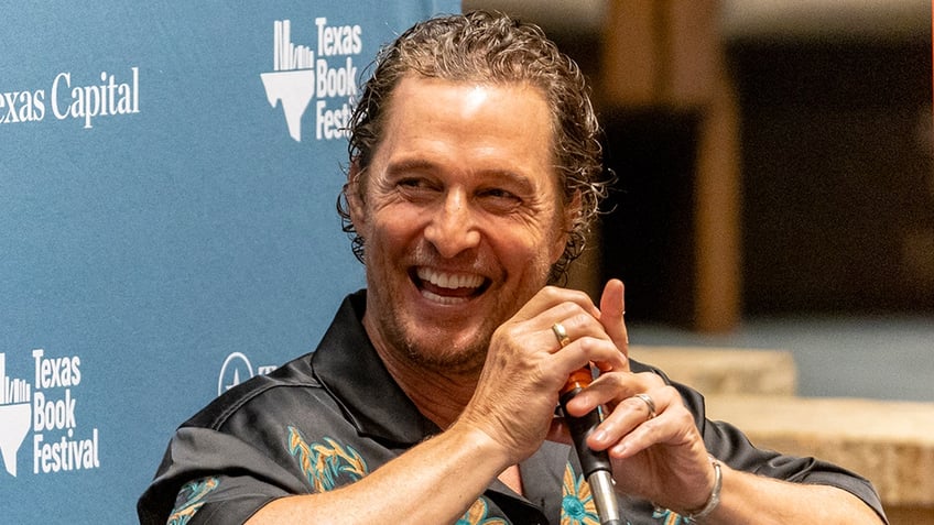 Matthew McConaughey laughs sitting back while wearing a black satin-like shirt with designs