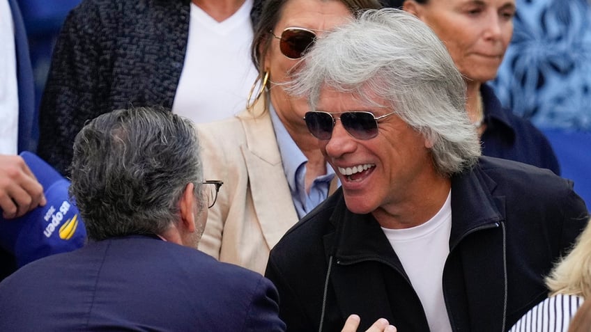 Jon Bon Jovi smiles as he greets someone at the US Open final wearing a black jacket and white t-shirt