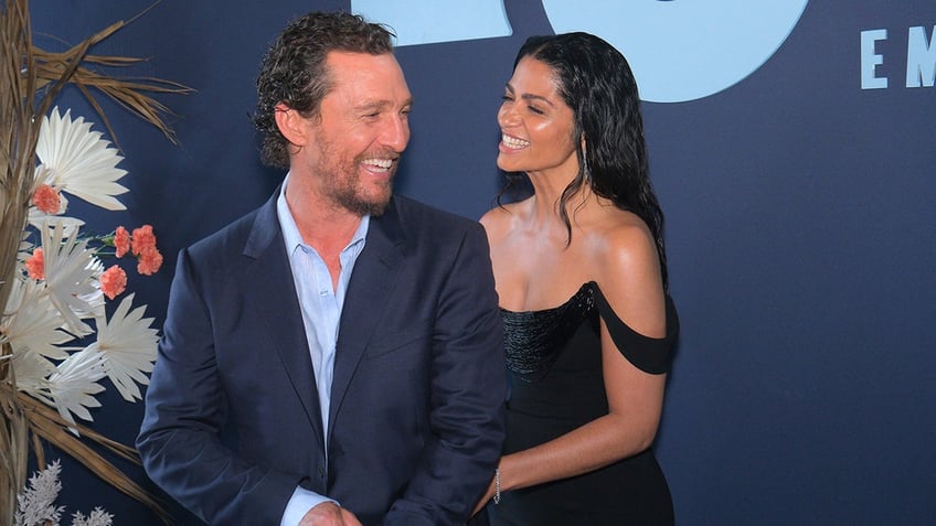 Matthew McConaughey in a blue shirt and navy blazer laughs as his wife Camila in a black off the shoulder dress