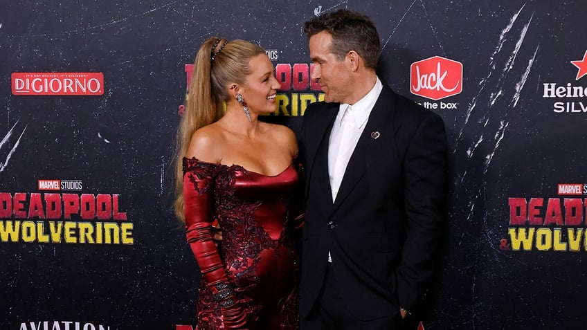 Blake Lively in a red jumpsuit looks lovingly at husband Ryan Reynolds in a black suit on the carpet