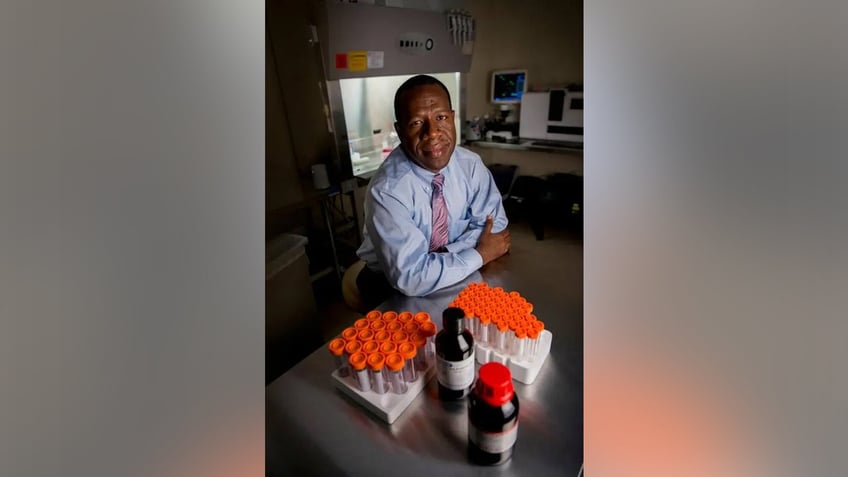 Dr. Zubair sits in his lab at a table behind two trays of test tubes with orange caps.