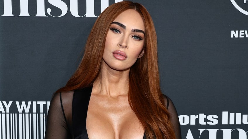 Megan Fox in a black plunging top with sheer sleeves on the carpet tilts her head