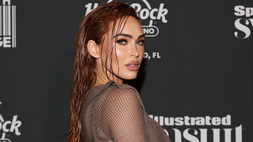 Megan Fox with wet-looking hair turns to the side in a mesh outfit and poses for the camera