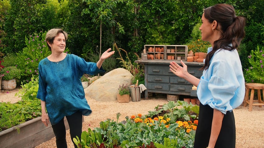 Meghan Markle wearing a light blue shirt in a garden opposite a woman in a bright blue blouse.