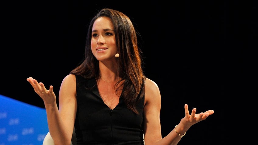 Meghan Markle with her arms raised wearing a sleeveless black dress and speaking to a mic.