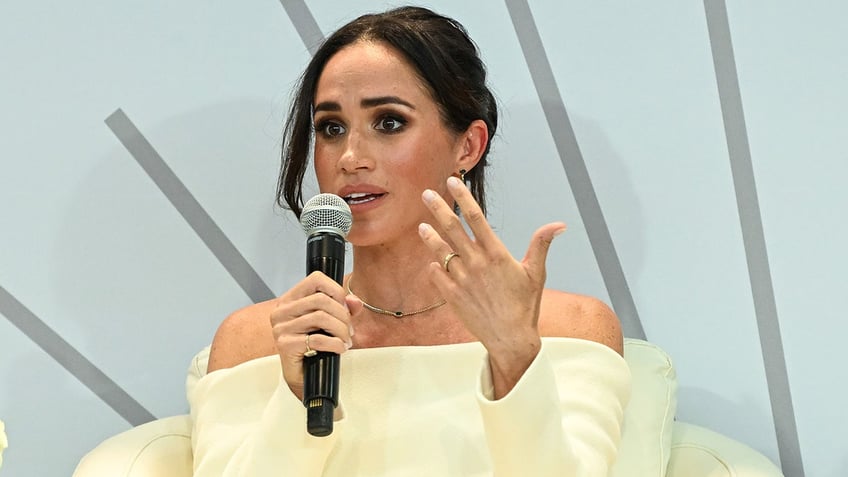 Meghan Markle wearing an off-the-shoulder beige sweater holding a mic and speaking to an audience.