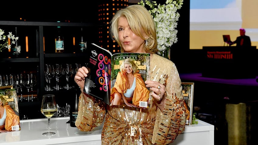 Martha Stewart in a gold dress blowing a kiss and holding her Sports Illustrated cover issue.