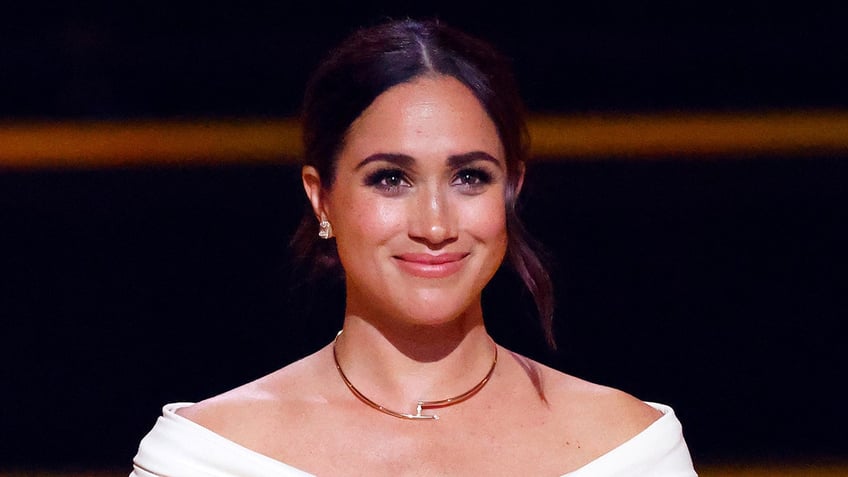 Meghan Markle smiling wearing a gold necklace and a white top.