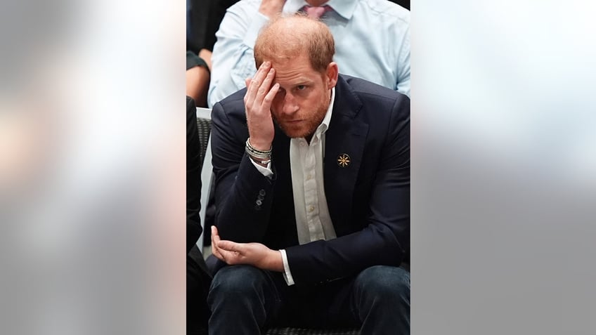 Prince Harry with his head down looking upset in a dark suit.