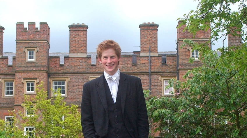 A young Prince Harry smiling in a traditional dark suit in front of Eton College.