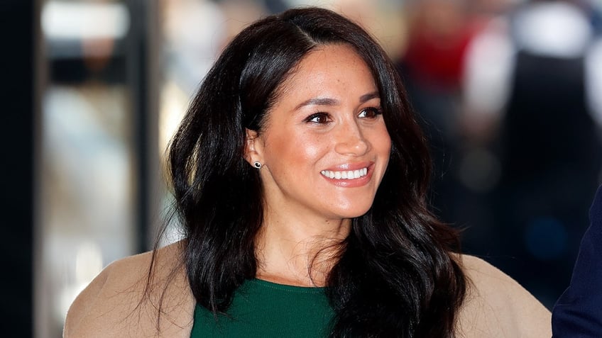 A close-up of Meghan Markle wearing a green dress and a beige coat as she smiles.