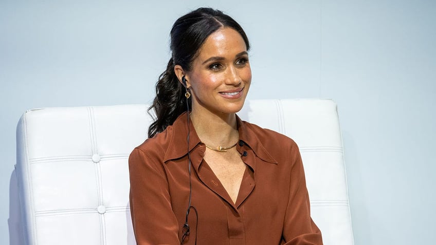 Meghan Markle wearing a brown blouse and sitting on a white couch.