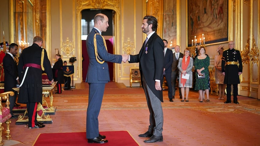 Prince William and Jason Knauf wearing suits inside the palace with people watching as the prince grants Knauf a prestigious honor.