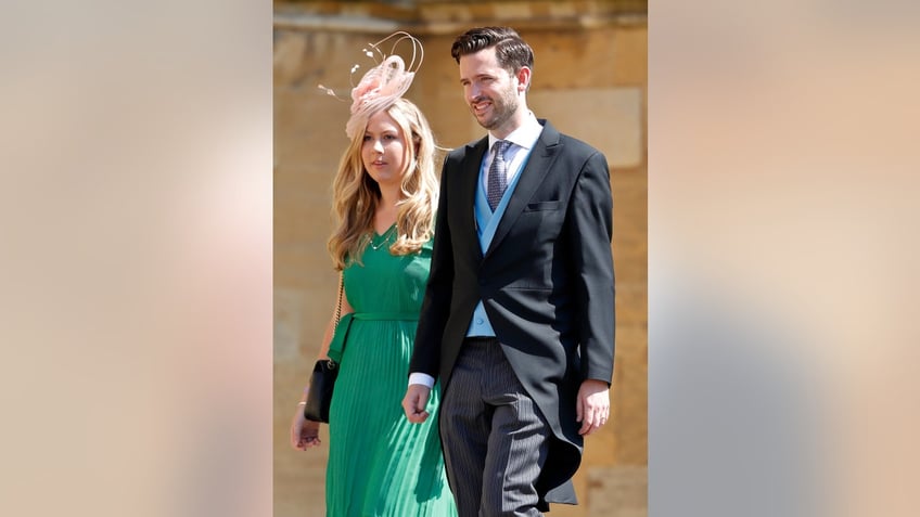 Jason Knauf wearing a suit and tie walking next to a woman wearing a bright green dress and a pink fascinator.