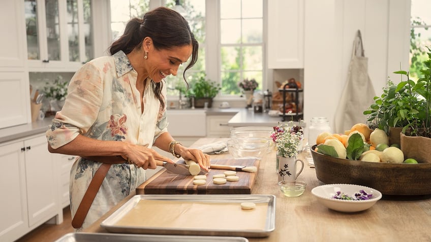 Meghan Markle smiling wearing a floral dress cooking in the kitchen.