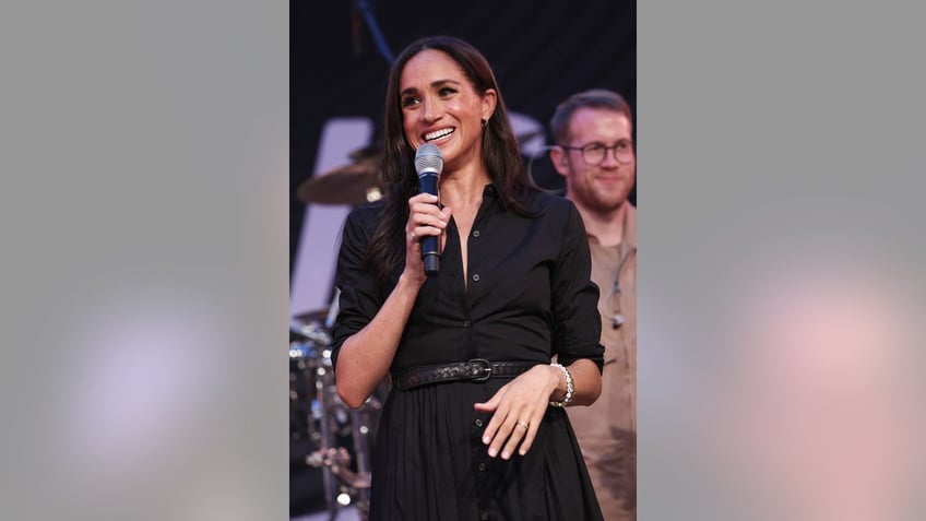 Meghan Markle smiling wearing a black dress and holding a mic.