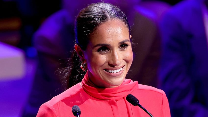 A close-up of Meghan Markle smiling in front of a mic wearing a red turtleneck.