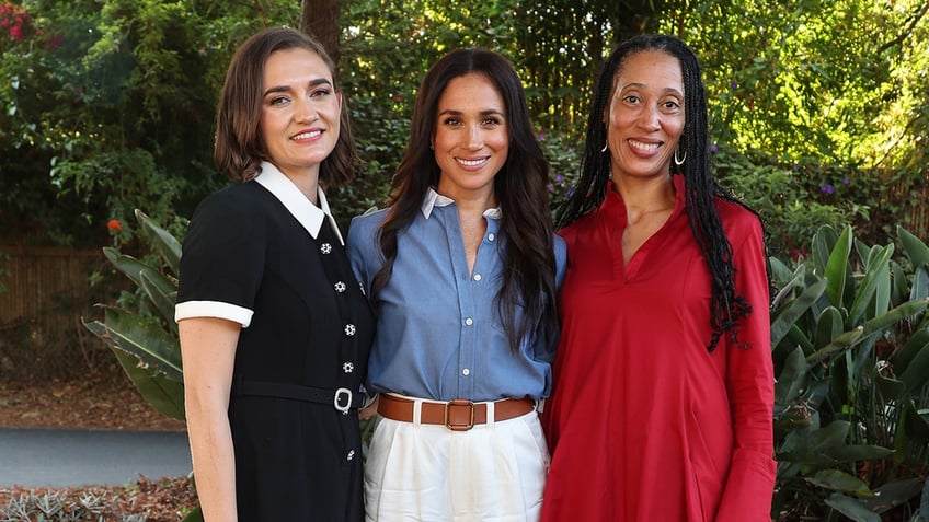 Meghan Markle wearing a blue blouse and white pants in between two women smiling