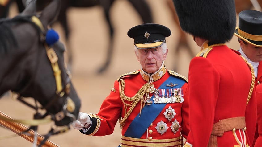 King Charles III in a red formal suit next to a horse