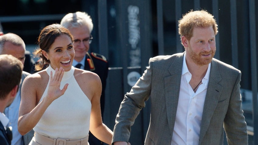 Meghan Markle waves in a white halter top and light pink pants while holding Prince Harry's hand in a grey suit