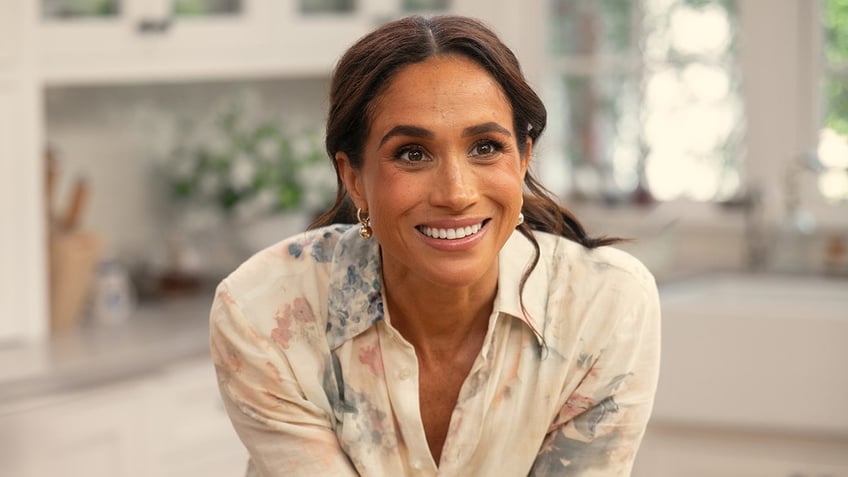 A close-up of Meghan Markle smiling and wearing a white and floral shirt dress.