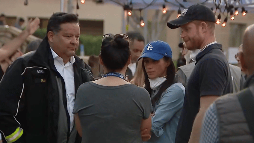 Victor Robo in a black jacket speaks with a woman with her back turned who is talking to Meghan Markle in a blue Dodgers hat and Prince Harry in Pasadena