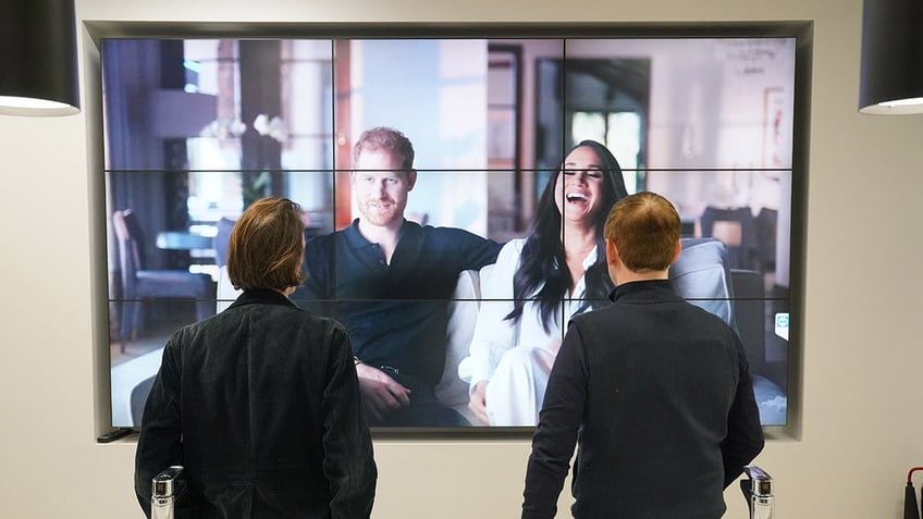 Two men watching Prince Harry and Meghan Markle on the big screen