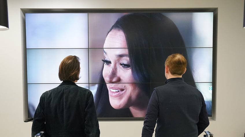 Two men looking at Meghan Markle on a big screen.