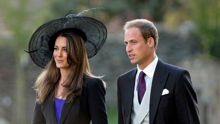 Kate Middleton wearing a blake blazer and a bright blue blouse with a black oversized hat standing next to Prince William in a suit and tie