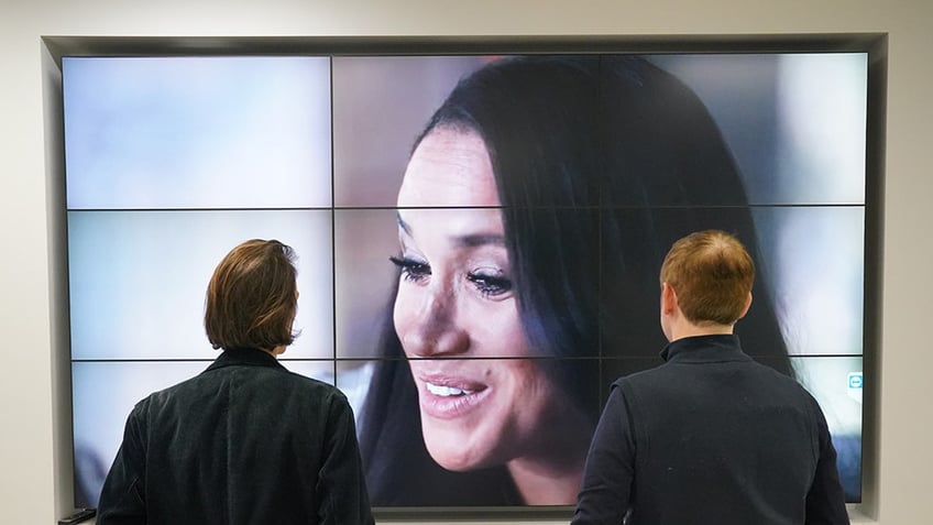 Two people with their backs turned watching Meghan Markle on television.