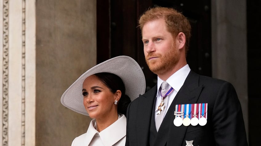 Prince Harry in a suit with medals standing next to Meghan Markle in a white coatdress with a matching hat.