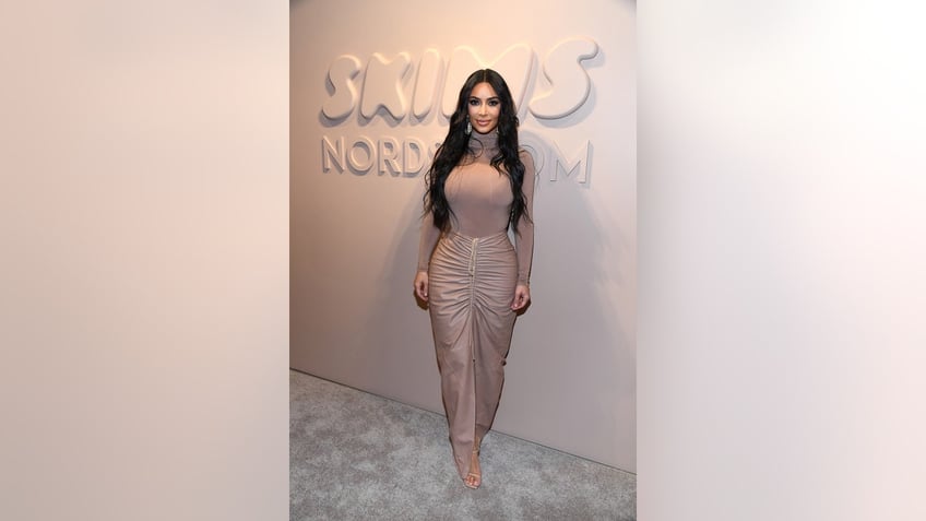 Kim Kardashian wearing a body hugging nude-hued outfit standing in front of the Skims Nordstrom logo