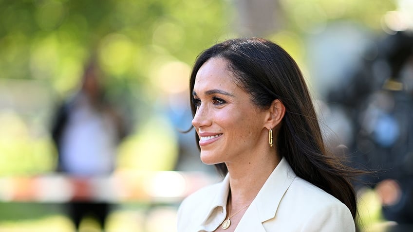 Meghan Markle smiling wearing a white blazer and looking to the side.