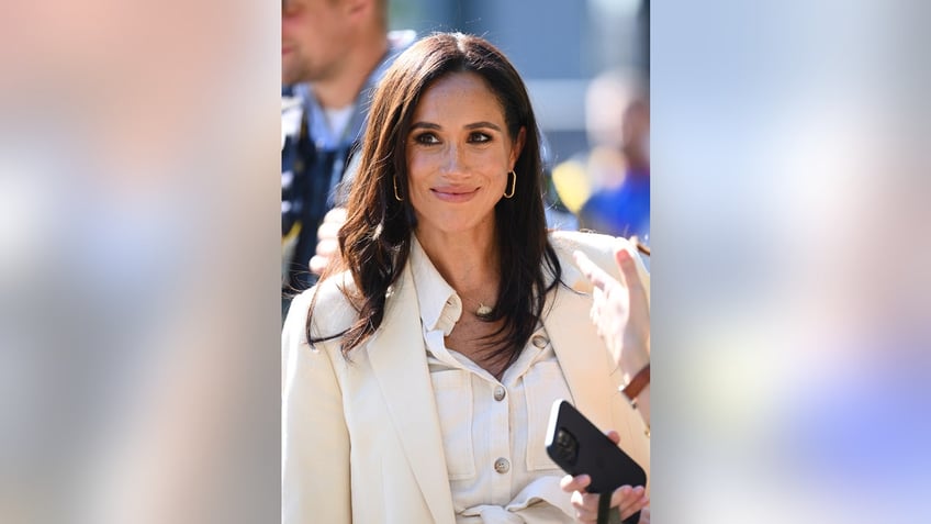 A close-up of Meghan Markle wearing an all beige ensemble and smirking outdoors.