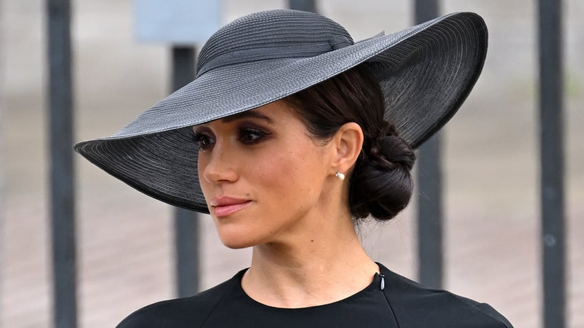 Meghan Markle looking to the side wearing a black dress and a black hat looking somber.