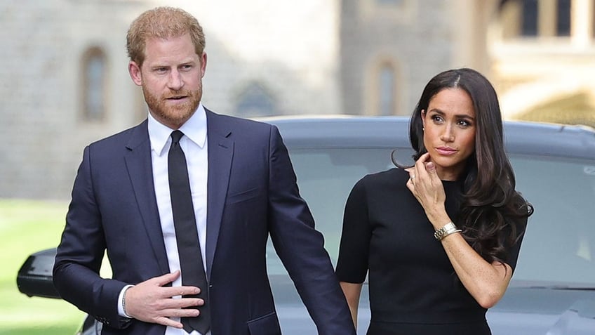 Prince Harry in a navy suit and tie holds Meghan Markle's hand, wearing a dark dress after the Queen passed away