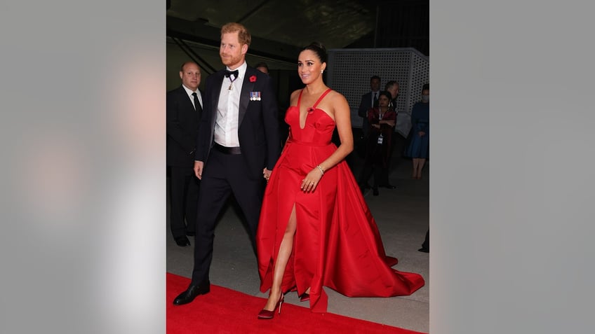 Meghan Markle wearing a red gown walking next to Prince Harry in a tux.