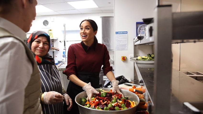 Meghan Markle wearing a burgundy sweater and gloves handling food