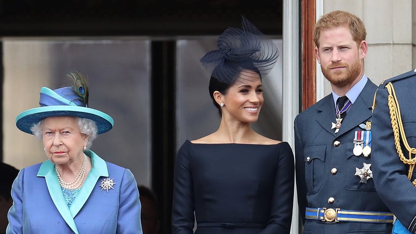 Queen Elizabeth wearing a blue and green coatdress standing next to Meghan Markle wearing a fitted blue dress and fascinator and Prince Harry wearing a suit with medals