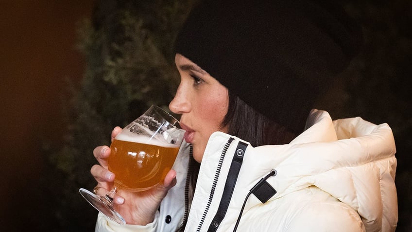 Meghan Markle drinking a beer from a glass while wearing a white coat and a hat.