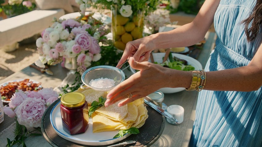 A close-up of Meghan Markle's hands next to a jar of jam and an array of plates.