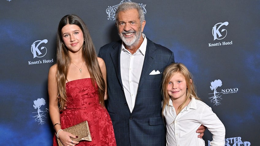 Mel Gibson in a blue suit poses on the carpet with his daughter Lucia in a red strapless dress and son Lars in a white button down and black pants