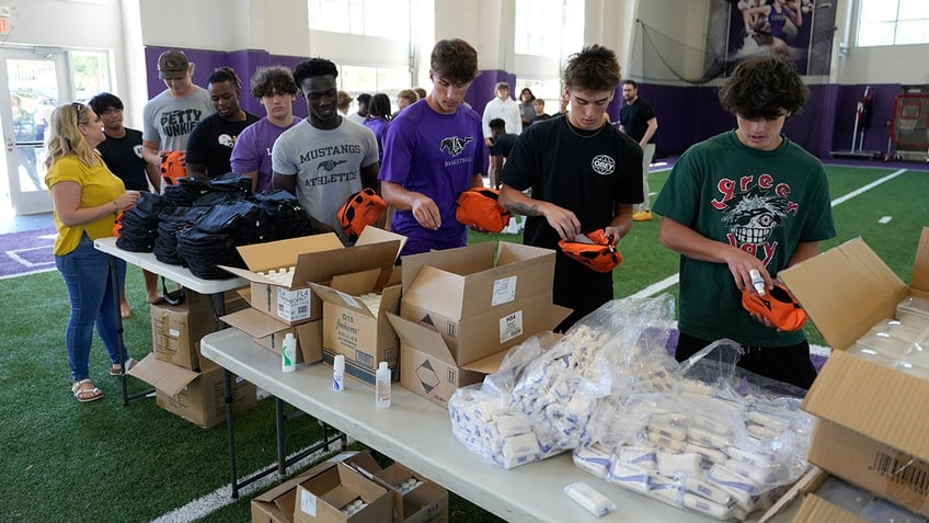 Melissa Joan Hart in a yellow shirt stands behind a group of teenaged boys filed in a line packing hygiene kits with World Vision