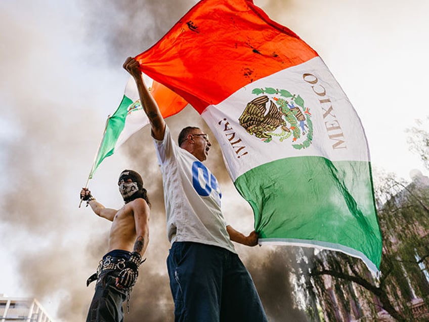 Two protesters wave Mexican flags while standing on a vandalized Waymo vehicle during a de