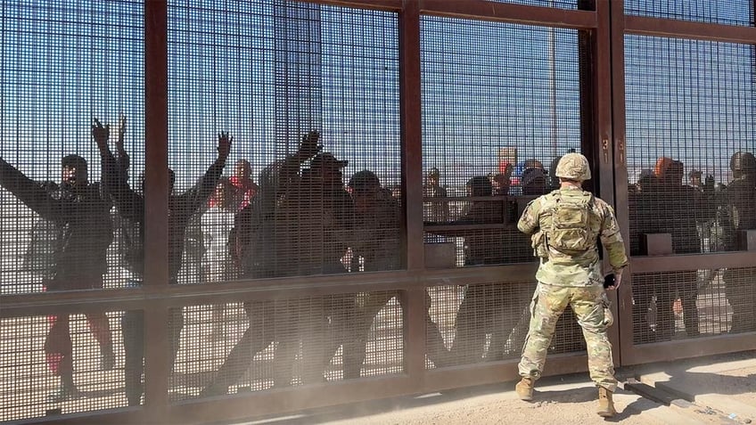 Migrants press up against border gate as Guardsman looks on