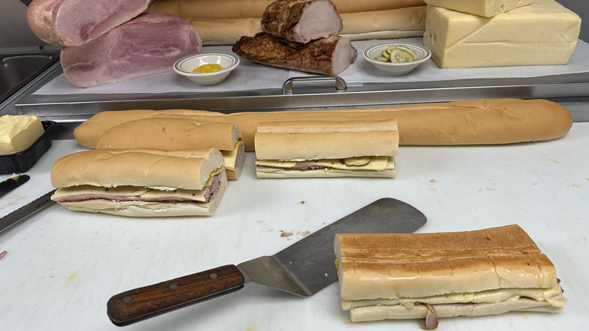 A freshly pressed Cuban sandwich from Versailles sits on a cutting board next to some unpressed sandwiches in the kitchen of the Miami restaurant, Sept. 26, 2024.