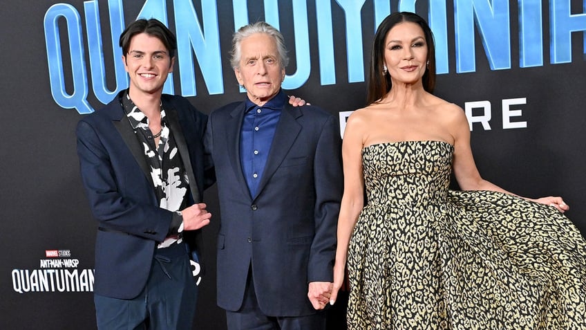 Michael Douglas in a navy suit and blue button down shirt poses with son Dylan also in a suit and wife Catherine Zeta-Jones in a leopard print dress on the carpet