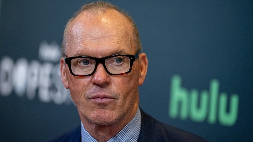 Michael Keaton in black framed glasses looks to his left seriously in a navy blazer and checkered blue shirt