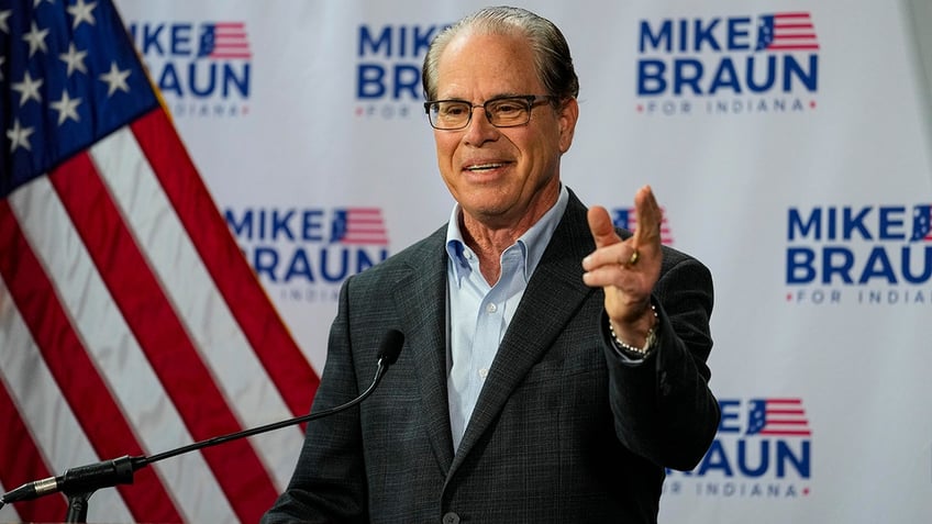 Sen. Mike Braun, gesturing with left hand in front of flag, campaign backdrop